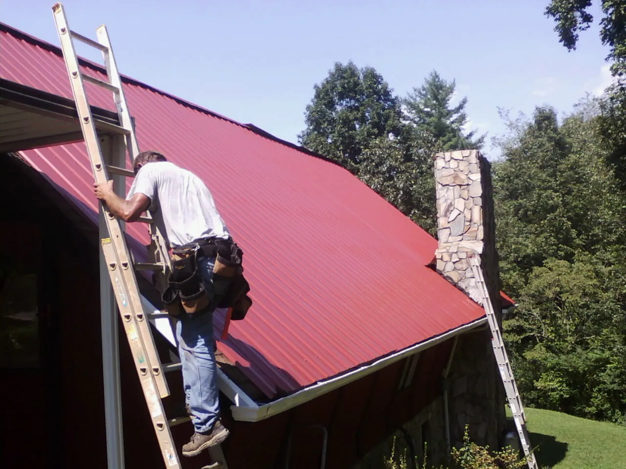 Roofer on a ladder beside a red metal roof and stone chimney — Soffit & Fascia Repair by Roofing Brothers in Lilburn